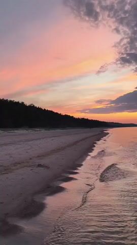 The video begins with a view of a sandy beach and the ocean at sunset. The sky is filled with pink and orange hues, and clouds are scattered across it. The waves gently lap at the shore. The camera then pans out to show a wider view of the ocean and the sky, with the sunset colors becoming more prominent. The video continues to show the ocean and sky, with the colors of the sunset shifting and fading as the sun dips below the horizon. The water is calm, and the reflections of the sky can be seen on its surface. The video ends with a shot of the ocean and the sky as twilight sets in.
