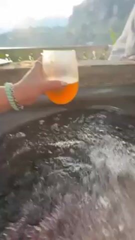 The video shows a person holding a glass of orange liquid in front of a bubbling hot tub. The camera then pans to reveal a scenic view of a lake and mountains, framed by a stone ledge with flowers and greenery. A second person is visible in the background. A small table sits nearby holding a speaker, a phone, and a framed picture. The overall impression is a relaxing setting with a beautiful view.