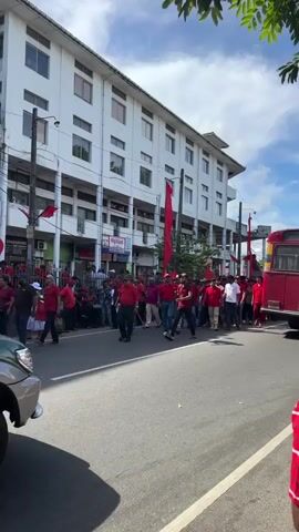 A street is shown in daytime, with a line of people dressed in red shirts walking along the street. Some of the people are carrying red flags on poles. A green Toyota Land Cruiser Prado is parked to the left of the street. Behind the people and the car is a white building with shops along the ground floor.  A scooter with two people aboard passes by on the street, moving opposite to the direction of the group.