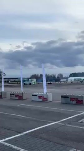 The video captures a dynamic scene at an outdoor driving event on an overcast day. In the background, a lime green truck is executing a drift, tires screeching and creating a cloud of white smoke. In the foreground, behind a row of gray concrete barriers labeled "FORMACJA FUN-DRIVE," a red passenger car also begins to drift, its tires kicking up dust. White flags with black text stand beside the barriers, adding to the organized feel of the event. Beyond the drift area, there are other vehicles, an open field, and structures in the distance, including what appears to be a warehouse or hangar. The ground is asphalt, marked with white lane lines, indicating a designated track or course.
