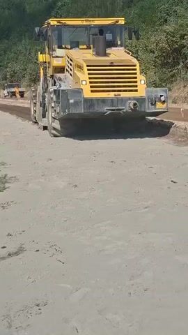 A yellow Bomag roller machine is shown compacting a dirt road. The machine is moving forward, with its large roller in contact with the ground. Trees and a bright blue sky are visible in the background. There are a few other vehicles and people in the distance on the road. The video appears to be taken from another vehicle moving alongside the roller.