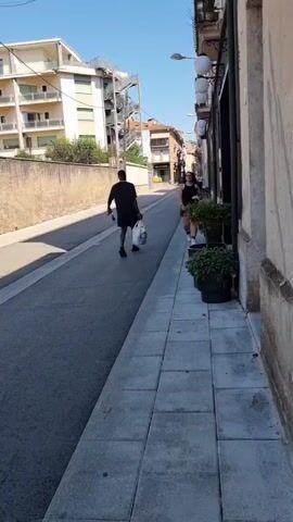 A man in a black t-shirt and shorts walks away from the camera down a paved street, carrying a white plastic bag in his right hand. A woman in a black tank top and a black skirt walks towards the camera on the sidewalk to the right of the street. She is carrying a white object in her right hand. She pauses and looks at the camera for a moment before continuing to walk. In the background, there are buildings and a blue sky.