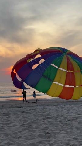 The video shows a beach scene at sunset. Two figures stand on the sandy beach near the water's edge, dwarfed by a large, multi-colored parachute. The parachute's design features a spectrum of colors including red, blue, green, yellow, and orange. Sunlight streams through openings in the parachute, highlighting its size and shape. The sky behind the parachute is filled with soft hues of orange, pink, and grey, indicative of the setting sun. The calm ocean reflects the warm colors of the sunset. Additional people are walking along the beach in the background. The overall scene is tranquil, emphasizing the natural beauty of the coastal environment and the scale of the parachute relative to the people on the beach.