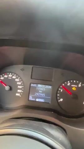 This media file shows a close-up of a car's dashboard and then a view from the driver's perspective while driving through a street.

The dashboard displays several pieces of information. On the left, a speedometer shows the needle pointing between 90 and 100 km/h. In the center, a digital display shows:
- "34.0°C" (temperature)
- "1156.5 km" (likely distance driven since last reset, or trip meter)
- "20:34" (time)
- "242965" (likely the total odometer reading)
- "70km/h" (current speed)
- A battery icon indicating a partially charged battery.

The second part of the media file shows the road ahead from the driver's seat. It appears to be a residential street with buildings on either side, characteristic of Boom, Belgium. The buildings are brick and have multiple windows. There are cars parked along the street and a few cars driving ahead. The sky is bright, suggesting it is daytime, possibly late afternoon or early evening given the displayed time of 20:34. The weather appears to be clear. The overall mood is mundane, depicting a typical drive.