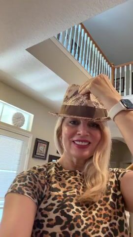A woman with blonde hair and red lipstick smiles at the camera while wearing a leopard print shirt and a brown patterned fedora hat. She adjusts the hat on her head with her right hand. The background shows a home interior with a staircase, railing, and a skylight.
