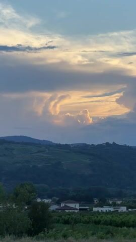 A wide shot captures a rural landscape under a cloudy sky during sunset. The sky is filled with dramatic, illuminated clouds in hues of yellow, orange, and blue, with some darker clouds present. In the middle ground, rolling hills are covered in dark green vegetation, possibly vineyards. In the foreground, there are some trees and a small cluster of buildings with red-tiled roofs. A white sign with green lettering "57.5" is visible on the right side of the frame. The overall scene is serene and picturesque.