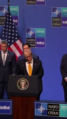 The video shows a man in a dark suit and yellow tie speaking at a podium. He is standing in front of a backdrop with NATO logos and flags. To his left, an American flag is visible. There are two other men behind him, also in suits. The man at the podium appears to be Donald Trump, based on his appearance. He is looking down at his notes and speaking into a microphone. The overall environment suggests a formal event, possibly a press conference or a summit.