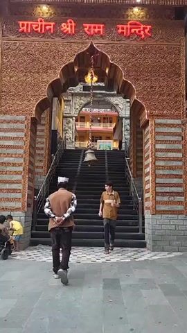 The video captures the entrance of the "Prachin Shri Ram Mandir" (Ancient Shri Ram Temple) in Manikaran, India. The temple features a grand, arched gateway adorned with intricate carved patterns, resembling terracotta or wood, and accented with brick-like stonework. A large, decorative bell hangs from the center of the archway, suspended by a chain. Above the arch, the temple's name is illuminated in bright red neon Hindi script.

Beyond the main arch, a wide, dark-colored staircase ascends into the temple complex. Another, simpler archway is visible at the top of these stairs, leading further into the temple grounds.

Throughout the video, several people are seen entering and exiting the temple. Two men are initially prominent in the foreground, walking towards the stairs; one wears a brown jacket and a white cap, the other a striped brown t-shirt. As the camera moves slightly, more visitors appear, including women in both traditional Indian attire (like kurtis) and casual Western clothing, along with a child playing nearby. People are steadily ascending and descending the stairs, engaging in typical temple visiting activities. The scene appears to be set during the daytime, although the neon lights on the entrance remain illuminated, adding to the visual appeal. The overall impression is that of a bustling yet peaceful religious site.