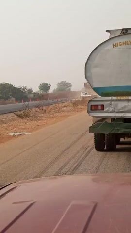 The video, taken from inside a vehicle, shows a white tanker truck with the words "HIGHLY INFLAMMABLE" written on the back. The truck is driving on a paved road, leaving dirt tracks. A white truck and a car pass the tanker truck in the opposite direction. The sky is hazy, and there are trees and orange traffic cones visible on the side of the road.