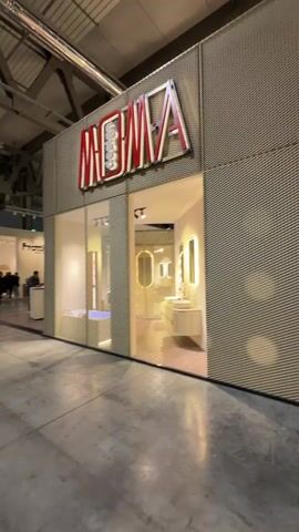 The video showcases an indoor exhibition hall, likely a trade show, in Rho, Italy. The camera initially focuses on a prominent booth with the brand name "MOMA" in large red and white letters, featuring bathroom fixtures inside. The booth exterior is a textured, possibly metal mesh material. The camera then pans left, revealing a wide exhibition corridor with a high, industrial-style ceiling. Other booths are visible, including one partially labeled "Frattini." Numerous people are seen walking along the corridor, browsing displays, and engaging with the exhibits. One person wears a black cap and uses a phone. The video then transitions into the interior of a brightly lit, modern booth with minimalist design, featuring light-colored walls, circular cutouts, contemporary lighting, and a curved white reception counter. Several individuals are present inside this booth, some standing and conversing, others walking around.