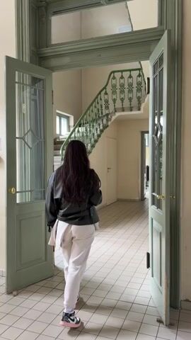 A person is seen from the back, walking from a room out into a hallway. The person is wearing a black leather jacket, white pants and black and pink shoes. They have long brown hair and a white bag over their shoulder. The hallway has a tiled floor and light-colored walls. To the left is a staircase with a green banister. The doorway that the person walks through has glass windows and is painted green.