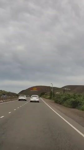 The video shows a gray sky with heavy clouds over a landscape. Two white cars are driving on a paved road. In the background, on a hill, there is a large symbol consisting of a yellow sun with rays and a red circle inside. There are also utility poles with wires visible along the side of the road and on the hill. The vegetation on the right side of the road is green and somewhat overgrown. The overall scene appears to be a highway leading towards a hillside monument under a cloudy sky.