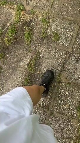A person wearing a white garment and black sneakers is walking on a stone tiled ground.  The person makes a peace sign with their hand. Some grass grows in between the tiles. A small piece of garbage is visible on the ground.
