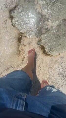 A first-person perspective video shows feet stepping into shallow pools of clear, mineral-rich water with a white, textured ground. The camera pans to reveal a wider landscape of white terraced pools with people bathing and walking around them. In the background, a town and mountains can be seen under a clear blue sky.