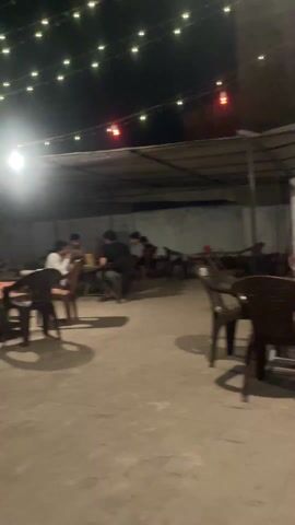 The video shows the outdoor seating area of a café or restaurant at night. String lights hang overhead, illuminating tables and chairs. Several people are seated at the tables, some appearing to be engaged in conversation. A man in a white shirt and dark pants is sitting in a wooden chair, looking to his right. The business name "SHAM KI CHAI" is visible on a wall in the background. Some people are sitting and standing.