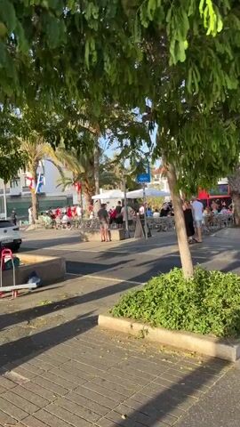 A public outdoor seating area at a cafe is depicted. A white car turns into the frame from the right, its driver visible. Several people are seated at tables under white umbrellas and palm trees. Some are wearing red and yellow flags, possibly indicating a sporting event. A man in the foreground is facing away from the camera, wearing a grey t-shirt and shorts. Flags are hung on poles and palm trees. The ground is paved with interlocking bricks. The time stamp indicates the video is 4 seconds long.