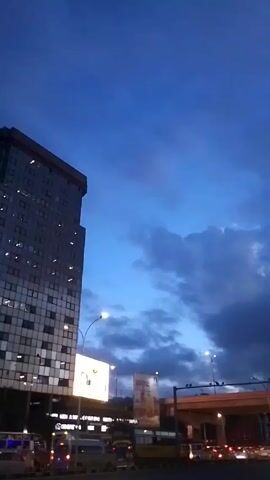 The video shows an urban scene at dusk. A tall, modern building with many windows stands on the left. The sky is a deep blue, with patches of gray clouds. Streetlights illuminate the scene, and a highway overpass is visible in the background. Traffic is present on the road, including vans and buses. Billboards are also visible near the overpass. The overall atmosphere is one of a city at the transition from day to night.