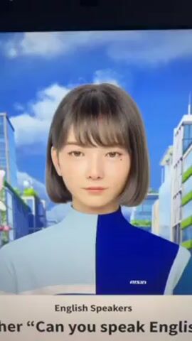 A close-up shot of a computer screen features a virtual female character with short, dark hair and bangs. She has a fair complexion and is wearing a two-toned outfit, light blue on the left and dark blue on the right, with "AISIN" printed on the right side. The background shows a stylized city scene with blue skies and fluffy clouds. Below the character, the text "English Speakers" is displayed, followed by the question "Can you speak Englis." The character's eyes shift, and her mouth opens slightly as if she is about to speak. The video appears to be a demonstration of AI technology or a virtual assistant. No other people or objects are visible in the shot.
