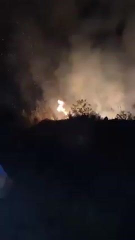 Here is a concise description of the video:

At night, a fire burns in a field, with flames visible behind a line of vegetation. Smoke and embers rise into the dark sky. The camera is static. Two voices can be heard talking about the fire in the audio.