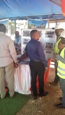 Here is a concise description of the video:

The video shows a group of four men under a blue and orange tent.  One man, dressed in a tan shirt and slacks, stands with his hands behind his back, facing left. Next to him, a man in a navy blue shirt and black pants is turned to face the right side of the frame.  He appears to be explaining a display board of photographs to two men in yellow safety vests and white hard hats.  A table draped with a white cloth and decorated with red fabric is positioned in front of the display board.  The ground under the tent is covered with gravel and loose rocks.  Another man wearing a blue patterned shirt and a red hat is visible behind the display board.