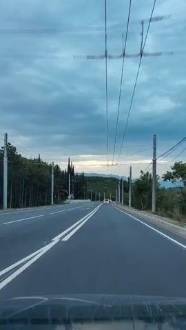 The video depicts a road as seen from inside a moving vehicle, likely a car. The road is asphalt, divided by double white lines indicating lanes for traffic. Overhead are several power lines stretching across the road. 

On either side of the road are patches of greenery including trees and shrubs. Notably, there are several tall, thin cypress-like trees lining the road. Utility poles are also visible along the sides. 

In the distance, there are hills or mountains covered in vegetation. A few vehicles are visible on the road ahead, their headlights on. The sky is overcast with dark, grey clouds, suggesting a gloomy or early morning atmosphere.