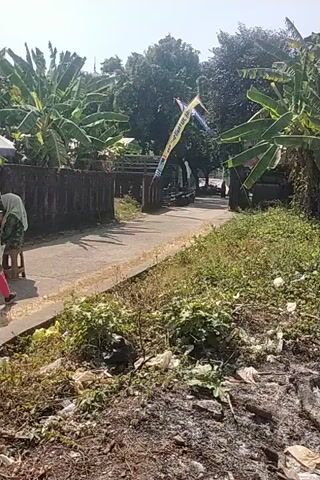 A video taken from a low angle, possibly from a moving vehicle, shows a street scene. The camera is tilted sideways. On the left, there are large green leaves, likely banana trees, and a brick wall. A tall, colorful kite, possibly a dragon kite, is visible in the distance. A pathway or road runs through the center of the frame, bordered by vegetation on the right.

In the foreground, on the pathway, a person in a yellow jacket and pink pants, with a backpack, walks away from the camera. Further down the path, under a large umbrella, are several people gathered around what appears to be a mobile food stall. Two children, one wearing a pink headscarf, are visible. One person is seated on a motorbike that is part of the food stall. A blue plastic stool is next to a bicycle. Another motorbike is parked in the foreground, obscuring part of the view. The ground is a paved pathway, with dry, bushy vegetation to the right. The overall lighting suggests a sunny day.