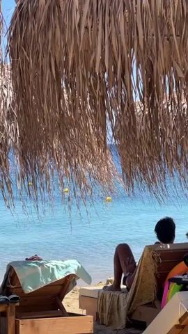 A beach scene with a thatched umbrella in the foreground, providing shade. Under the umbrella, beach chairs are visible. One chair is occupied by a person viewed from behind, wearing dark clothing and sitting with their legs extended. Another chair next to it is empty and covered with a blue and white striped towel and some other items. In the background, the turquoise ocean meets the shore. Several yellow buoys are floating in the water. A few people are visible in the water, one appearing to be a child in a pink swimsuit. The sand is light brown. The lighting suggests a sunny day.