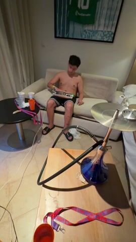 A young man is sitting shirtless on a light-colored couch in what appears to be a hotel room, playing a handheld game. He is wearing black shorts and black sandals. A hookah sits on a small wooden table in front of him. There are two small, round tables on either side of the couch. One table holds several empty white and orange cups and a pink lanyard. The other table has a silver disk, possibly part of the hookah setup, and some white objects. A red and purple lanyard is on the floor near the hookah table. A framed image of a green soccer jersey hangs on the wall behind the couch. The room has beige tiled floors and light beige walls.