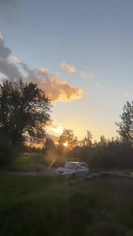 The video shows a view from a moving vehicle of the landscape. In the distance, there is a forest edge and a sunset. The clouds are illuminated by the setting sun, and the trees are silhouetted against the sky. The foreground is a green, grassy area. The vehicle is moving at a moderate speed.