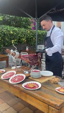 A man in a white shirt and black apron with a logo is slicing a large ham on a silver stand. He is wearing black gloves and is using a long, thin knife. The ham is dark red with streaks of fat. Several plates of thinly sliced ham are arranged on a wooden table in front of the ham. There are also small breadsticks on one of the plates. The scene appears to be outdoors, with greenery and a black umbrella in the background.