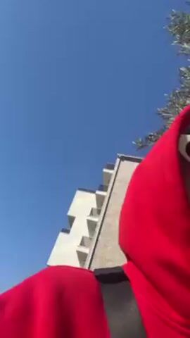 A person wearing a red hooded sweatshirt with a word in Cyrillic script on the front and white rectangular sunglasses walks along a path with a fence to the side. The person has a black shoulder bag. Trees and buildings are visible in the background. The sky is blue and the sun is shining. The video appears to be handheld and the camera angle is slightly canted.