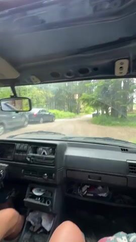 The video is shot from inside a car, looking forward through the windshield. The car's interior is visible, including the dashboard, steering wheel, and seats. A person's legs are visible in the foreground on the right side of the frame. The car is on a dirt road surrounded by trees and greenery. In the distance, there are houses and other cars. The car appears to be in motion, with the road and surroundings blurring slightly. The lighting suggests it is daytime.