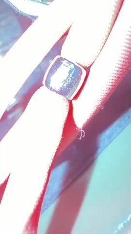 This is a close-up, static shot of a shiny, metallic bead clamped onto two strands of red, textured cord. The cord appears to be elastic, possibly part of a drawstring or bungee cord. The background is out of focus, showing a dark, muted color that could be fabric or a container. There are no people or any discernible action taking place. The lighting casts a strong reflection on the bead, highlighting its smooth, polished surface.