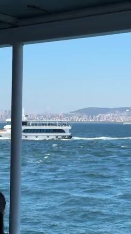This media file depicts a scenic view from a ferry on a bright, sunny day, likely in the late morning or early afternoon, given the clear blue sky and strong sunlight. The primary subjects are the sparkling blue sea, the distant cityscape, and a passing ferry.

The foreground is partially framed by the overhang of the ferry the viewer is on, with a white vertical support post visible on the left. The sea is a vibrant blue, with gentle waves creating whitecaps that catch the light. A white ferry with dark-tinted windows is in motion, moving from left to right across the frame, leaving a wake in its path.

In the background, a sprawling cityscape rises from the water's edge, nestled against rolling hills. The buildings are densely packed, with many appearing to be white or light-colored, interspersed with taller structures and what appear to be minarets, suggesting a location with Islamic architecture. The hazy atmosphere slightly softens the details of the distant city.

The overall mood is serene and picturesque, evoking a sense of travel and exploration. The clear weather and bright sunshine contribute to a feeling of pleasantness and relaxation. The presence of the ferries and the bustling cityscape indicates a populated area, likely a major city in Turkey given the "Prince Islands, Turkey" context. No visible text is present in the media file.