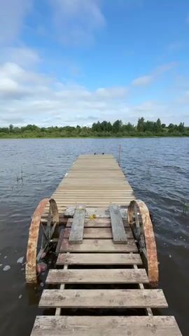 The video shows a wooden pier extending into a body of water. The pier is equipped with two large, rusted wheels on either side at the near end. Beyond the pier, the water extends to a distant shoreline lined with trees. The sky above is blue with scattered white clouds. The scene is calm and appears to be in a natural setting. There are no people or animals visible in the frame. The perspective is from a low angle, looking straight down the length of the pier.