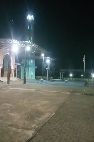 The video displays a nighttime scene of a mosque and its surrounding area. A tall minaret, illuminated with green lights, stands on the left side of the frame. In the center, a mosque structure is visible, also lit by bright lights. The ground in the foreground is paved with concrete, and a portion of a brick walkway is visible on the right. The overall ambiance is dark, with artificial lighting highlighting the architectural elements of the mosque.