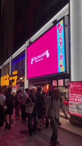 The video shows a bustling street scene in Japan at night. Pedestrians walk along a tiled sidewalk past shops and restaurants. A large digital billboard displays colorful imagery and advertisements, including one for "Churasun6 Okinawa" and another for "Happy Gaku Day." The scene is well-lit with artificial lights from the shops and billboard. People are dressed in casual clothing and appear to be going about their evening activities.