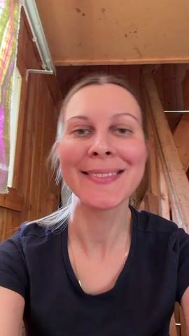 The video shows a woman centered in the frame, wearing a dark blue t-shirt and a delicate gold necklace. She is positioned within a wooden structure, possibly an attic or a loft, with unfinished wooden planks visible behind her and to the sides. She is looking directly at the camera with a smile, then puckers her lips, looks upwards and to the side before returning to a softer smile. The lighting is somewhat dim, but sufficient to discern details. A reflective, iridescent object hangs partially visible in the upper left corner of the frame.