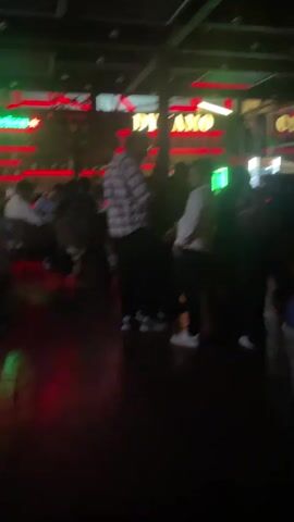 The video shows the interior of a dimly lit bar or club.  A sign that reads "DYNAMO" is visible in neon lighting above a bar area where several people are gathered.  There are bottles displayed behind the bar.  In the foreground, several individuals are standing and interacting.  To the left, there are tables and chairs where other patrons are seated.  The overall atmosphere appears to be social and lively, with patrons engaged in conversation.  In one scene, a couple appears to be posing for a photo in front of a fan. The flooring is wooden. Red and green neon lights are prominent throughout the space.