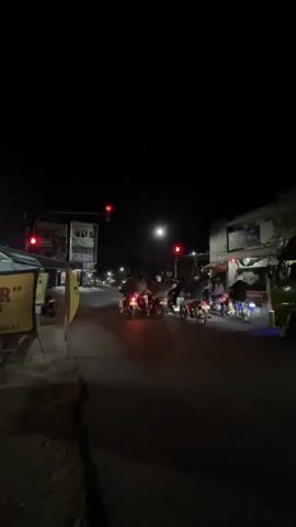 The video shows a street scene at night. Traffic lights are visible, cycling between red and green. Several motorcycles are on the road, some with riders. A yellow banner with text in Indonesian is displayed on the left side of the street. Buildings line the right side of the street. The overall impression is of a typical urban street at night with active traffic.