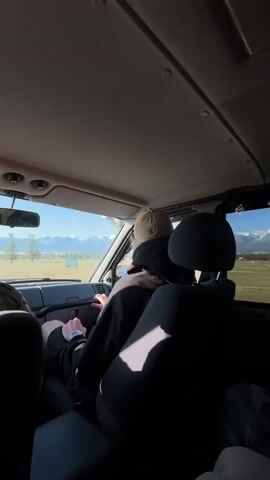 A first-person perspective video taken from inside a vehicle shows the view through the windshield and side windows. The camera faces forward, capturing a person in the driver's seat wearing a black hoodie and a tan baseball cap, their back to the camera. Their hands are on the steering wheel. The driver's side door is partially visible on the left. The vehicle's interior, including the ceiling, dashboard, and the back of a headrest, are prominent in the foreground and middle ground. Sunlight streams in through the windshield, creating highlights and shadows within the vehicle. Outside, a vast, flat landscape stretches towards a range of snow-capped mountains in the distance under a clear blue sky. The terrain appears to be a mix of open fields and scattered trees. The video appears to be shot while the vehicle is in motion, suggesting a road trip or travel through a scenic area.