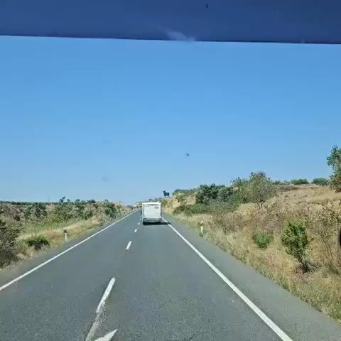 The video shows a view from inside a vehicle looking out at a road. A white camper van is traveling on the road ahead. To the right of the road, on a hill, there is a large, dark silhouette of a bull. The sky is clear and blue, and the landscape is dry and grassy with some scattered trees and bushes. The overall scene appears to be a roadside view in a rural area on a sunny day.