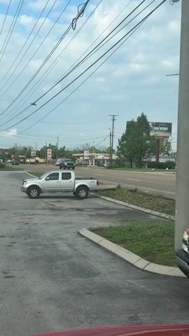 The video shows a street scene, possibly a parking lot and a road. A silver pickup truck is parked on the left side of the frame. A large gray SUV is driving on the road in the distance. Further down the road, there are some buildings and a billboard. A red pickup truck is parked in the foreground on the right, with part of its hood visible. The video appears to be filmed from inside a vehicle, looking out through the windshield and side mirrors. The sky is cloudy.
