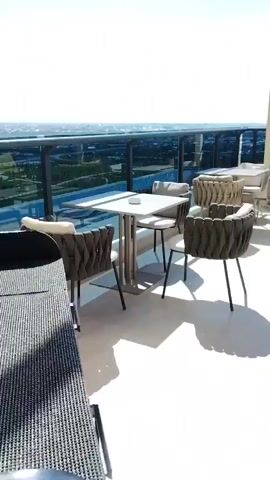 The video shows an outdoor patio or rooftop seating area with several tables and chairs. The furniture appears to be modern and made of woven material with cushions. In the background, there is a railing overlooking a body of water and a cityscape or landscape. The sky is clear and blue, suggesting a sunny day. The area is empty of people, giving it a serene and vacant appearance.