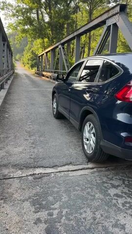 A dark blue SUV is parked on a narrow, weathered bridge. The bridge has metal trusses and a guardrail on the left side. The road surface is cracked asphalt. Trees line the sides of the road leading away from the bridge, and a river flows beneath it. Sunlight filters through the trees. The video pans across the river and the surrounding landscape, showing a valley with rolling hills in the distance. Ducks are visible in the water. The sun is low in the sky, casting long shadows.