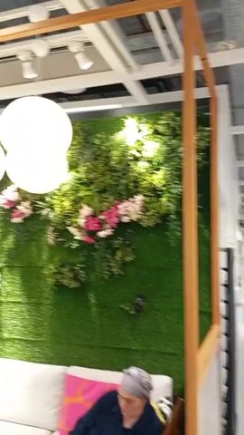 The video opens with a view of a green, artificial plant wall adorned with pink and white flowers. Several large, round, white paper lanterns hang from the ceiling in front of the plant wall. The camera then pans to the left, revealing a seating area with white sofas and cushions. To the left of the plant wall is a wooden structure with slats. Further left, a glimpse of a retail store interior is visible with shoppers and displays. The camera then moves down and to the right, focusing on a woman with dark, curly hair wearing a black top. She is bending over a small wooden table where a vase of white flowers and a bottle of water are placed. In the background, people are seen browsing in the store, some pushing strollers. The overall environment appears to be a section of a retail store, possibly a furniture or home goods store, designed to resemble an outdoor or garden-like setting.