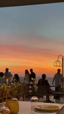 People are dining at an outdoor restaurant during sunset. The sky transitions from orange to pink and blue. Some people are taking pictures. There are tables set with glasses and cutlery. A couple is standing and embracing. A lamp illuminates a corner of the frame. The restaurant overlooks the sea.