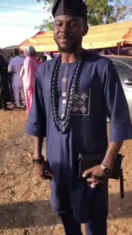 A medium shot captures a smiling man outdoors. He is dressed in a navy blue traditional outfit with a matching black hat, a triple-strand black beaded necklace, and a black beaded bracelet on his left wrist. He holds a black clutch in his left hand and wears a watch on his right wrist. Behind him, several individuals mingle near a beige tent or canopy. A silver car is also visible to the right. The ground is covered with brown dirt or mulch. The sky is light blue.