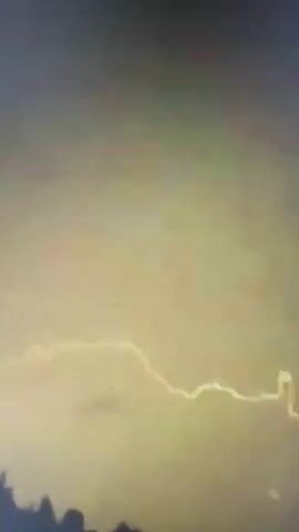 The video shows a cloudy sky with lightning strikes. The initial frames are dark and indistinct, then transition to a sky with an orange and pink hue. A bright yellow lightning bolt then flashes across the screen, illuminating the dark clouds. The lightning strike appears to be a jagged line extending horizontally across the frame. The video ends with the lightning strike and clouds becoming darker.