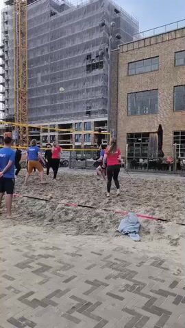 This is a daytime, wide shot video of a beach volleyball game being played in an urban setting.  In the background, a tall building under construction is visible, covered in scaffolding and a white protective sheet. To the right of this building is a brick building with large windows.  The volleyball court itself is made of sand, with a yellow net stretched across the middle.  Several people are playing volleyball.  On the left side of the court, closer to the viewer, a person wearing a blue t-shirt and dark shorts stands facing away from the camera.  Across the net, on the right side, several players are actively engaged in the game.  One person in a red shirt is jumping to hit the ball. Another person in a blue shirt is also near the net.  In the foreground, a paved area with a brick pattern separates the viewer from the sand court. A piece of light blue clothing is lying on the sand near the right sideline.
